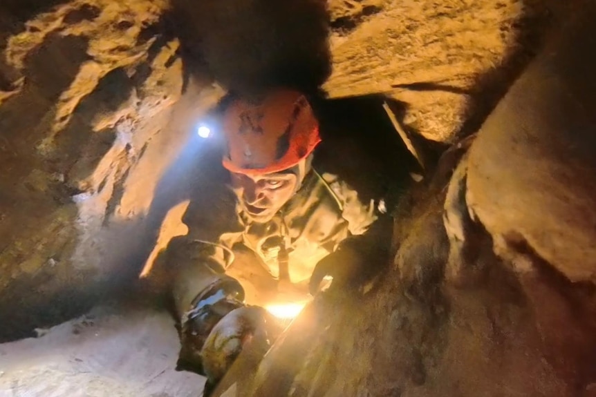 A man in a suit and orange helmet climbing backwards in a cave.