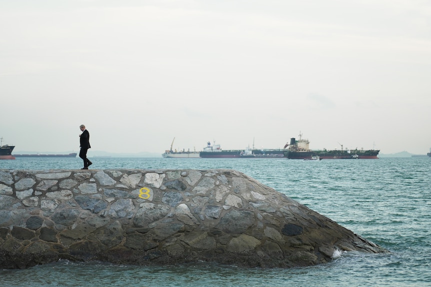 A man in suit walks on a groin at the bay in Singapore, a tanker ship in the background