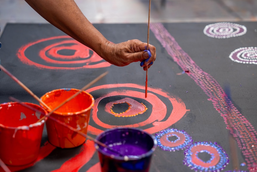 A close-up of some colourful paint pots and a hand painting on a black canvas with red swirls of colour. 