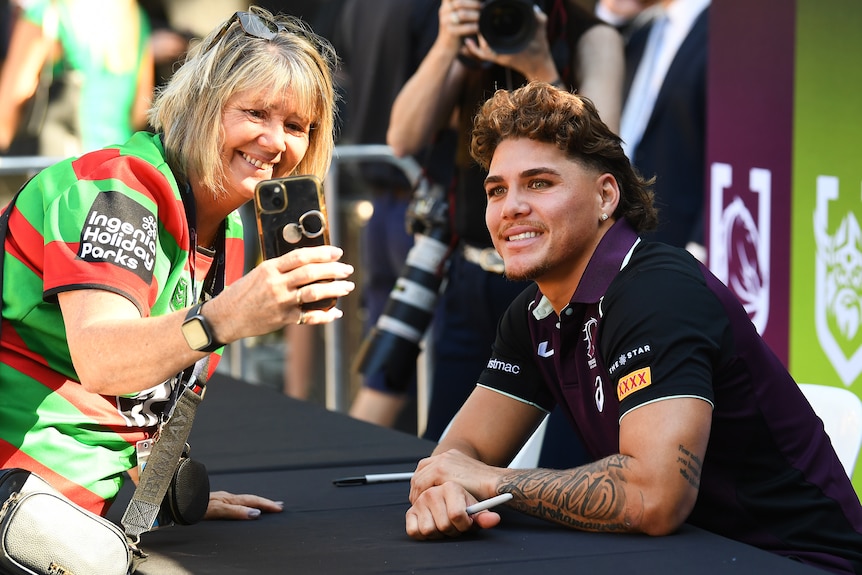Brisbane NRL player Reece Walsh sits behind a desk and smiles as a South Sydney fan holds her phone to take a selfie.