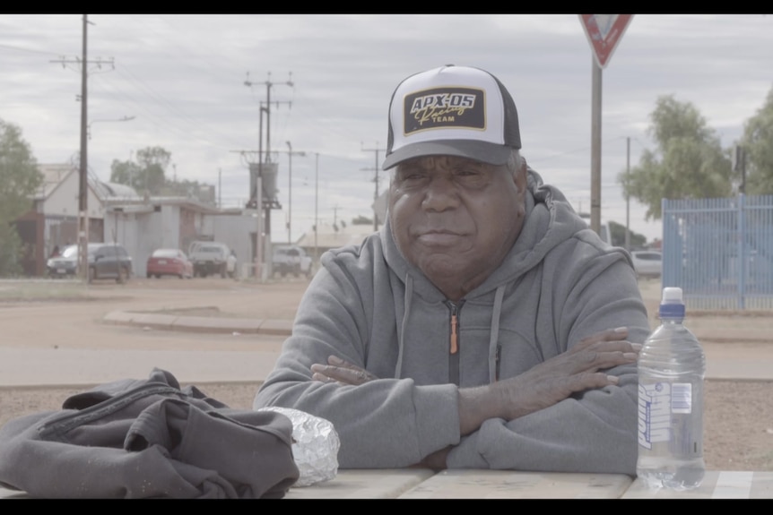 Man sits at table with arms crossed wearing cap