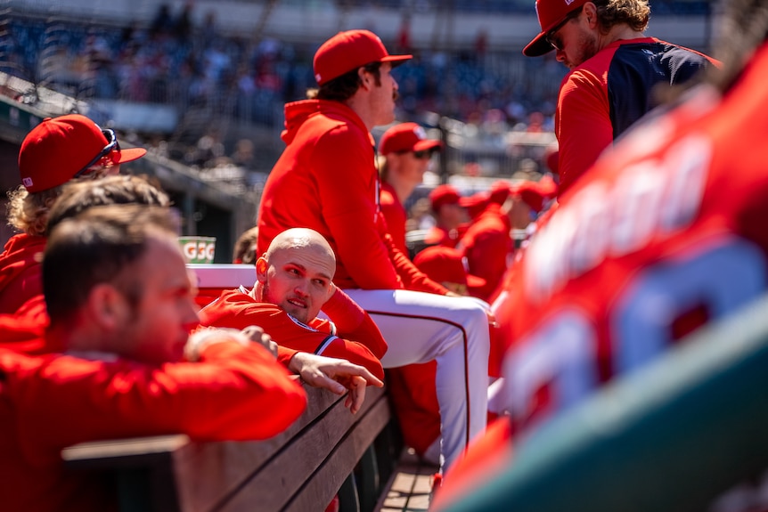 Curtis Mead, wearing red kit, leans on a barrier surrounded by his baseball teammates in the bleachers.