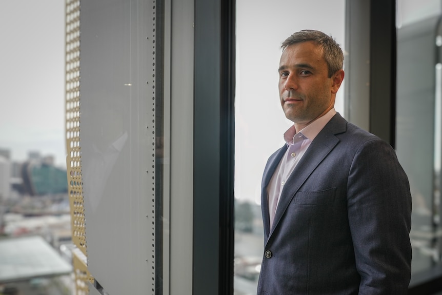 Chris Brycki in front of a window, wears a suit and open shirt.