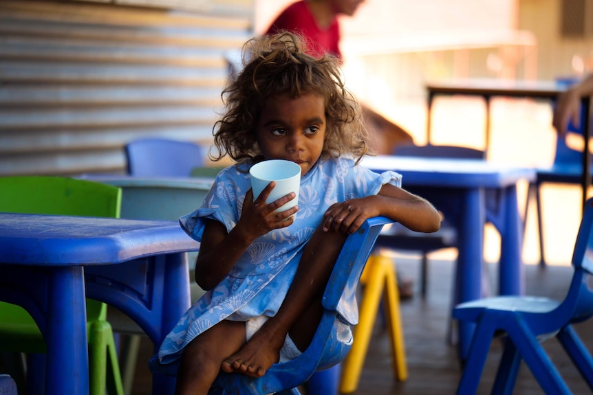 A young Aboriginal girl stares into space in thought, chewing on the rim of a plastic cup.