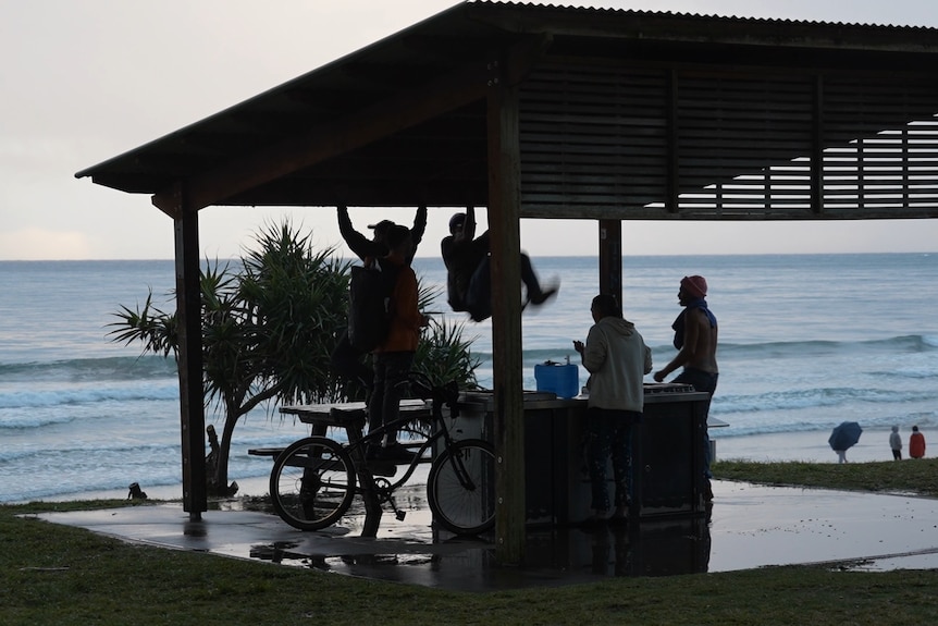 A beachside barbecue, table and youths hanging off the shelter and a bike, beach in the background