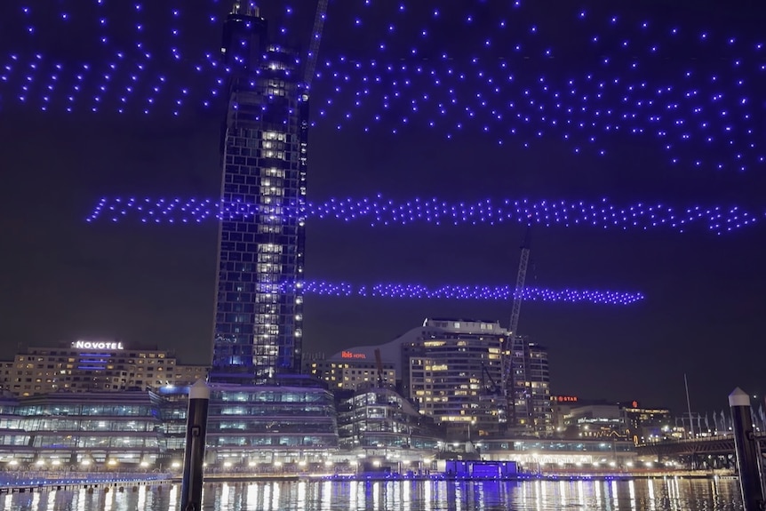 Drone blue lights in horizontal lines, a skyscraper in the background.