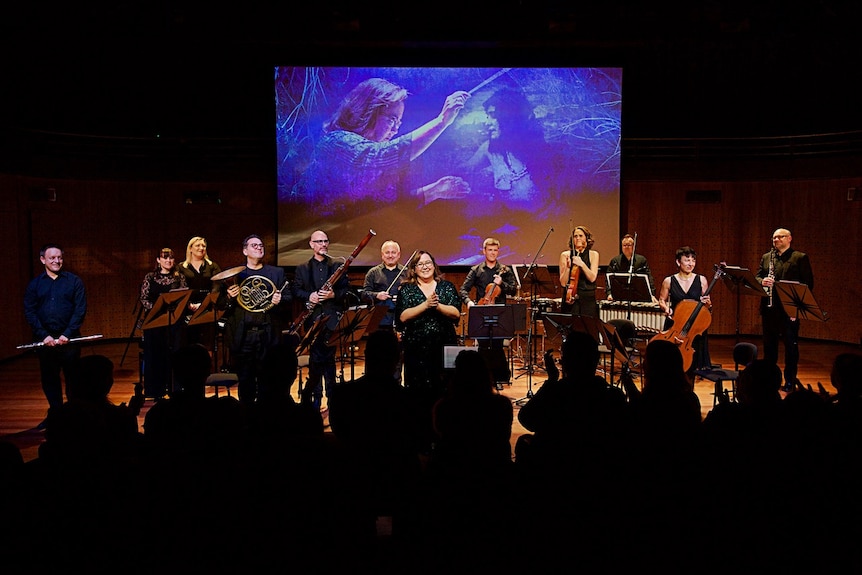 Jessica Wells and a chamber ensemble on a concert stage take a bow, with audience silhouettes and a blue-lit image on screen.