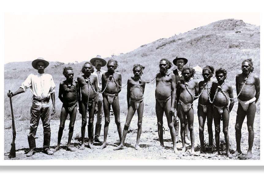 An old photograph of Aboriginal men wearing chains around their necks, flanked by armed guards.