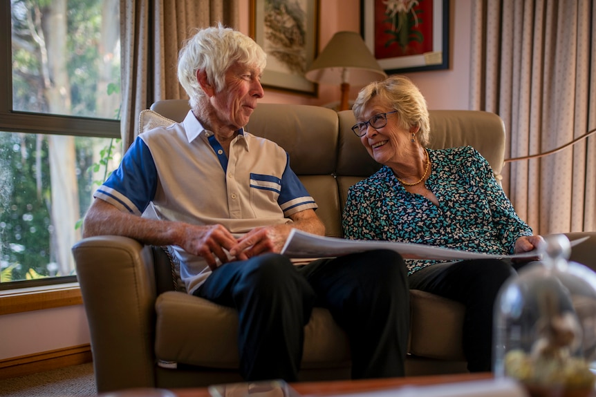 A couple smile at one another on a leather couch with greenery through windows.