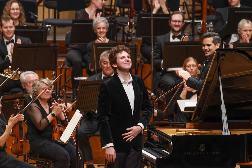 Jayson Gillham takes a bow standing next to a piano while orchestra members behind him applause