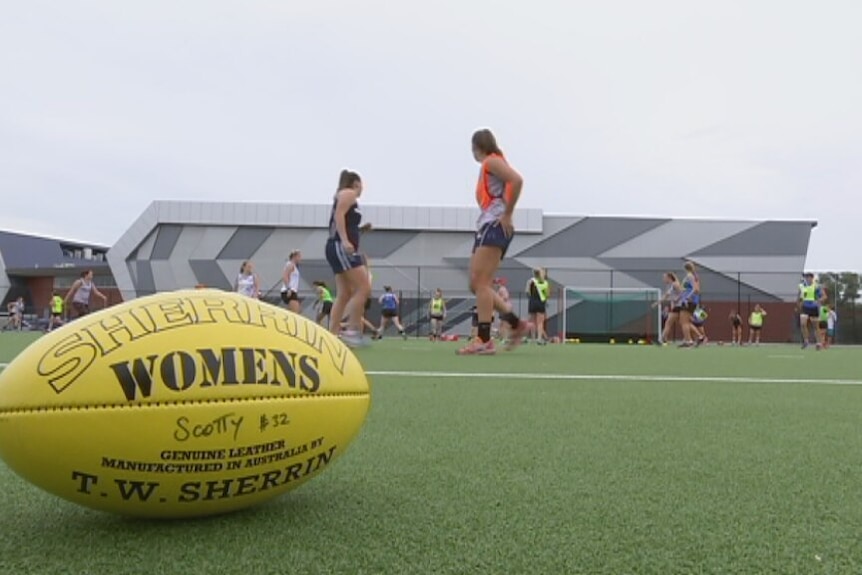 a yellow afl football on a grass field. Women run in the background.