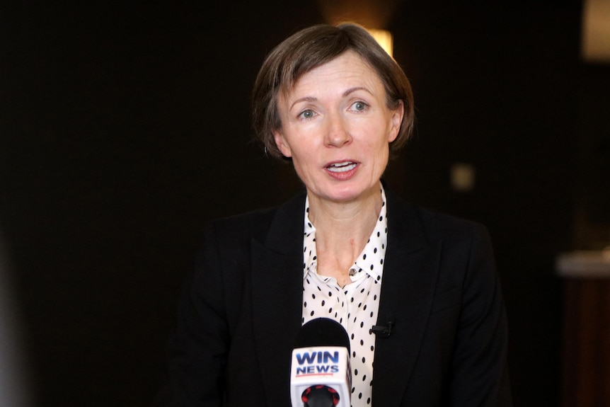 Woman in a dark jacket and white shirt talking into a microphone.