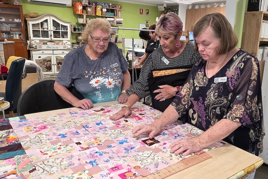Three ladies look down at a patchwork quilt.