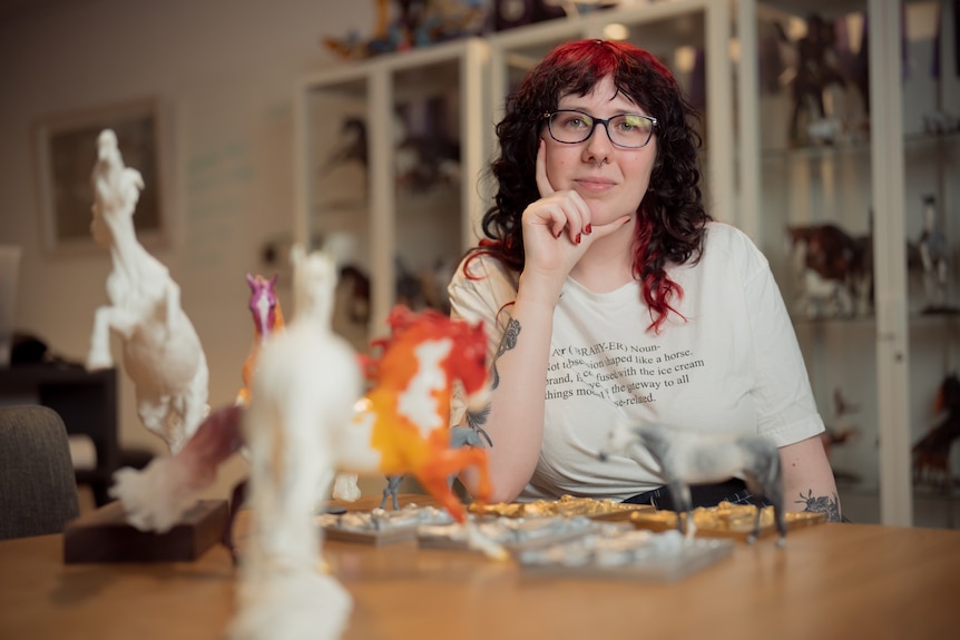 Maeve King in white shirt with glasses and red hair with model horses on table