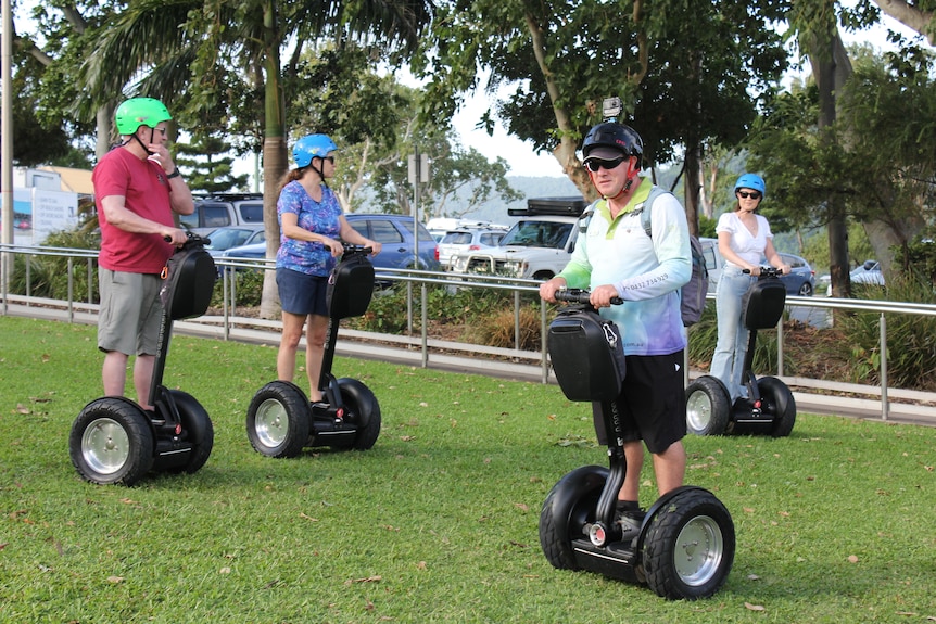 Four people ride on two-wheeled segways on a patch of grass outside a sailing club.