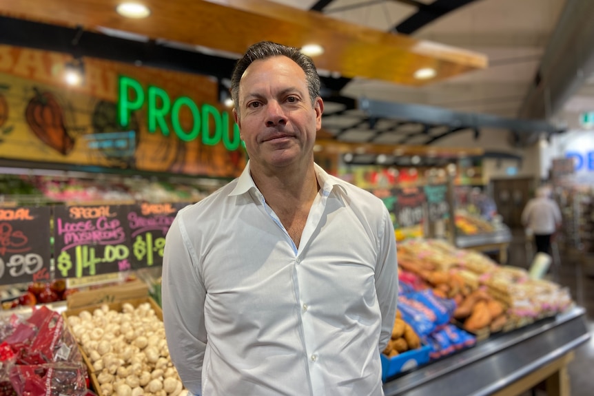 a photo of a man in white shirt standing in front of vegetables in supermarket