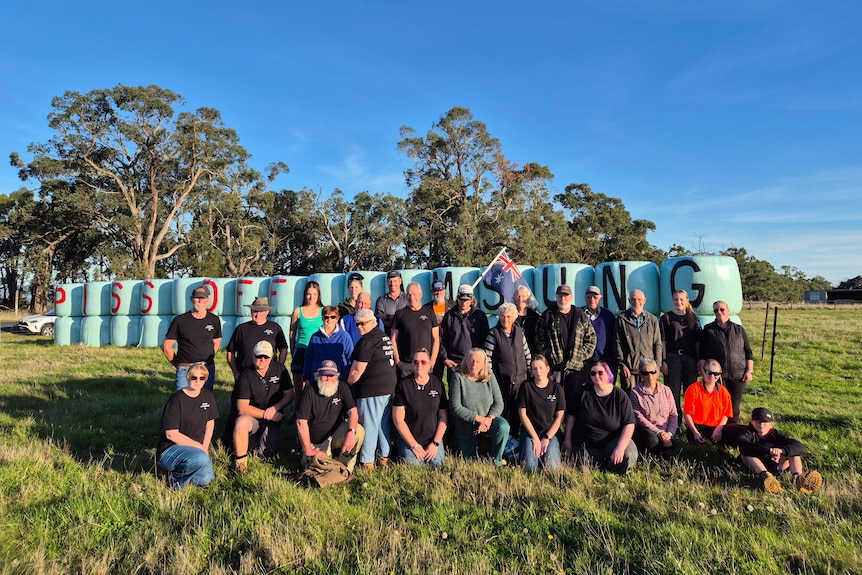 A group of people, many in black tshirts, stand in front of plastic-wrapped silage bales that say Piss Off Samsung