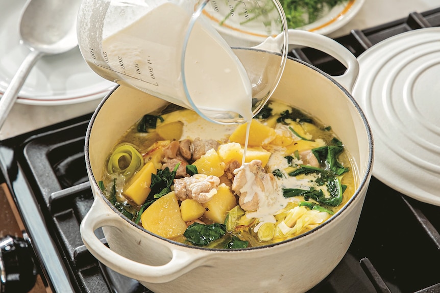 Cream being poured into a chicken stew simmering in a beige dutch oven on a gas stovetop.