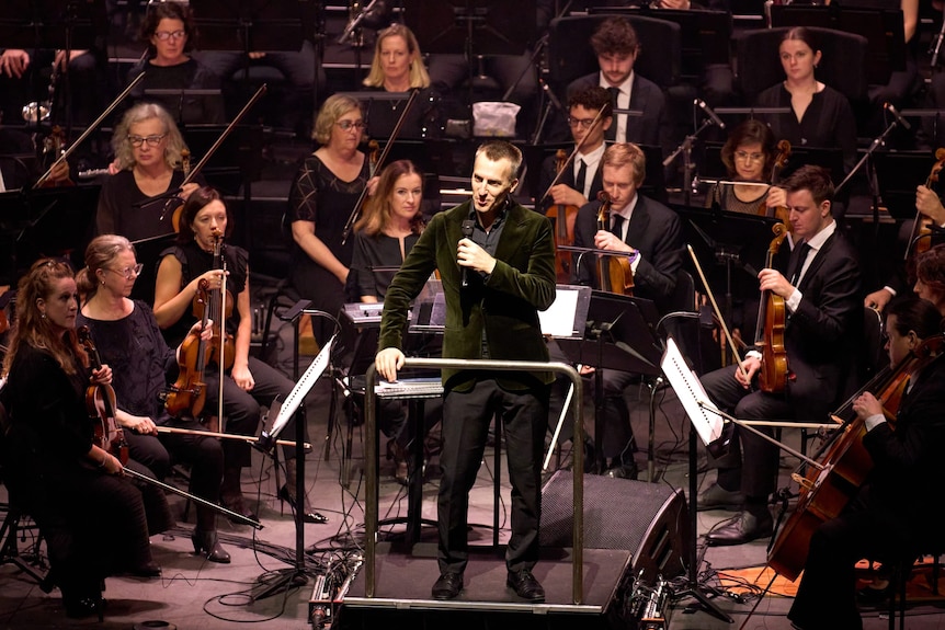 Nicholas Buc addresses the audience from the conductor podium, surrounded by string players, music stands and stage lighting.