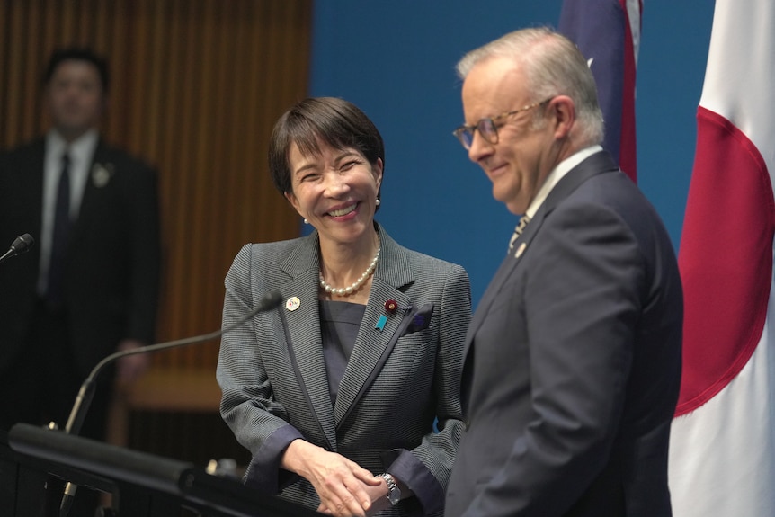 A man and a woman talk at a lectern with flags behind them