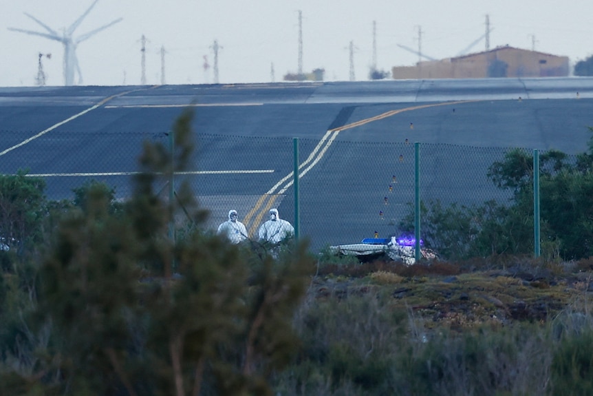 Two health workers in full biohazard get-up stand on the tarmac of an airport.