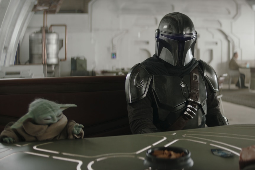 A little green creature and a man in armor sit at a table together