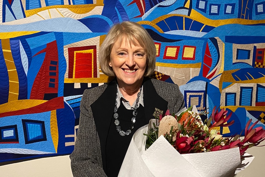 Older lady smiles while holding flowers and standing in front of a vivid handmade quilt.