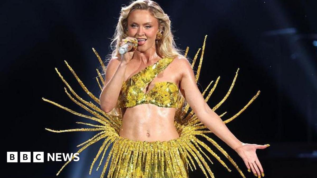 Zara Larsson wearing a yellow sparkly two piece outfit. She has long blonde hair and she is singing into a microphone.