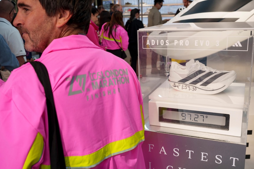A man in a bright pink jacket that says London Marathon stands next to an Adidas shoe in a glass cabinet.