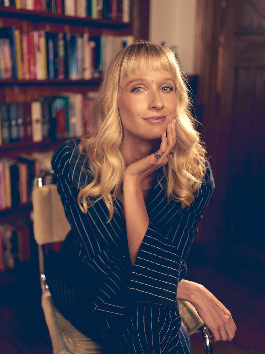 Dee Salmin sits leaning forward looking at the camera in a blue pinstripe suit with a bookshelf behind her