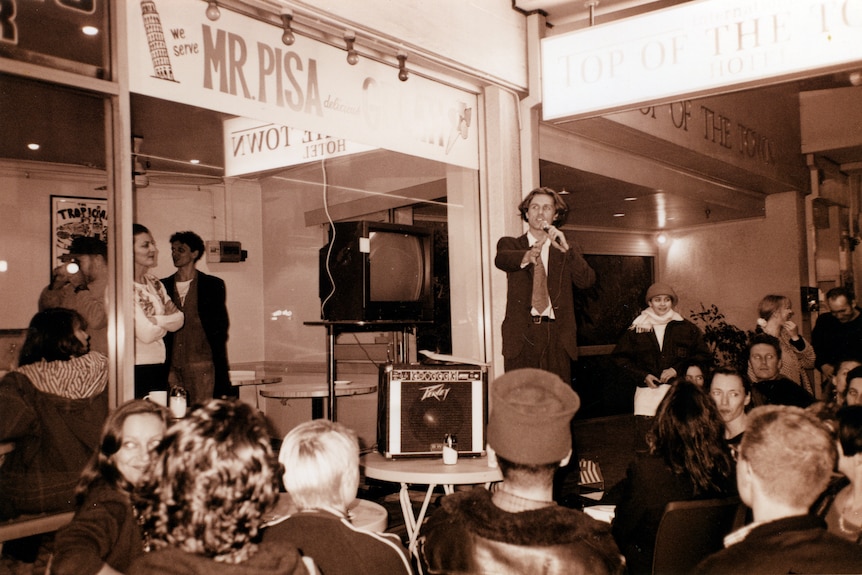 Black/white imagery, man in a loose-fitting suit stands on a chair to address a crowd. Next to him is a speaker and analogue TV