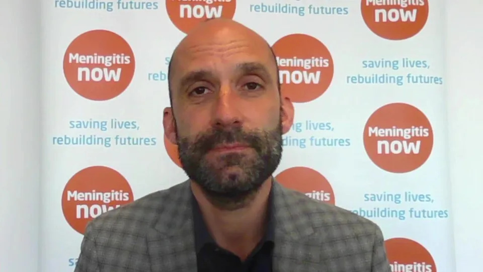 Dr Tom Nutt, chief executive of Meningitis Now, sat in front of a banner promoting the charity. He is a balding man with facial hair, a checked blazer and black shirt.