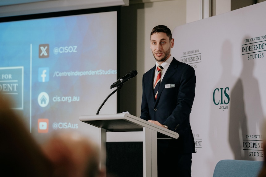 An adult man standing up at a lectern speaking to a crowd