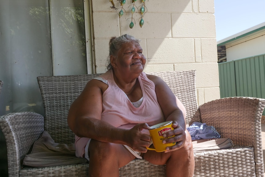 An older, grey-haired woman sits with a large mug of tea on a couch out the front of her house.