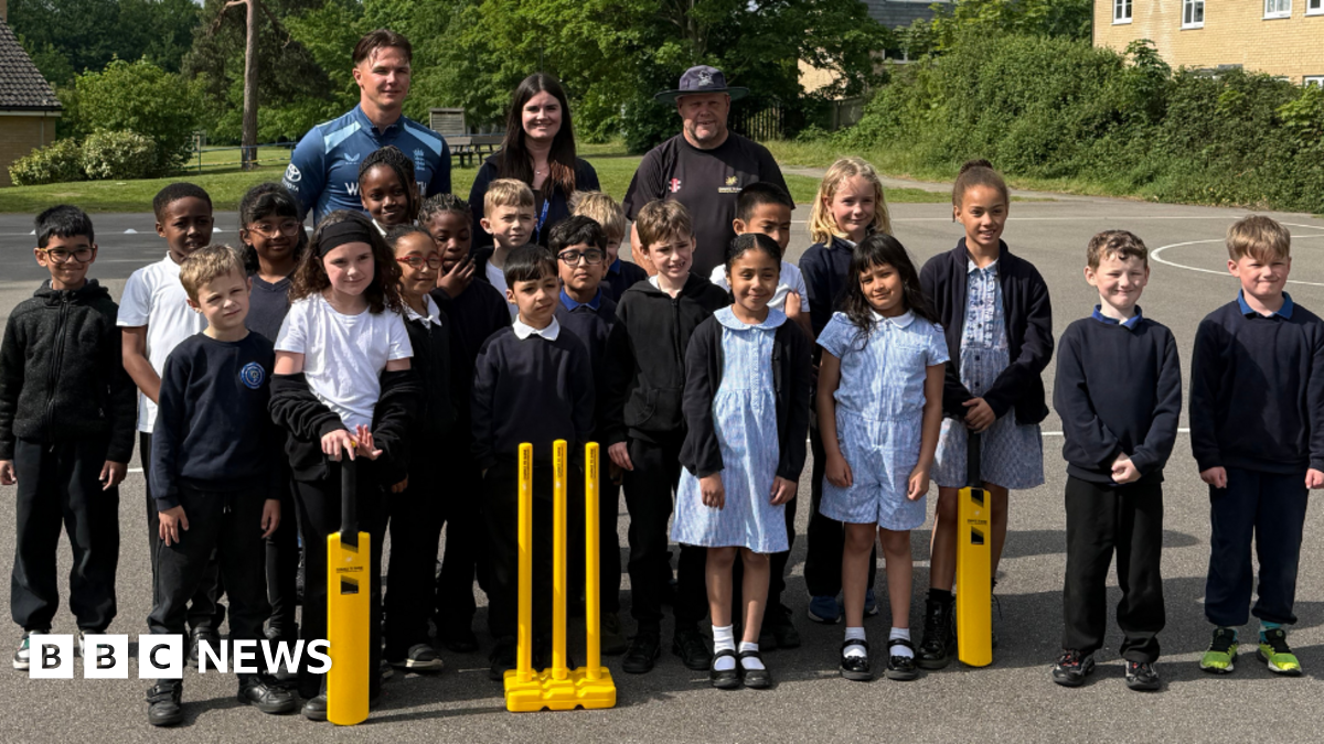 A group of school children standing together in front of yellow wickets and a bat. Behind them are three adults, including English fast bowler Sonny Baker.