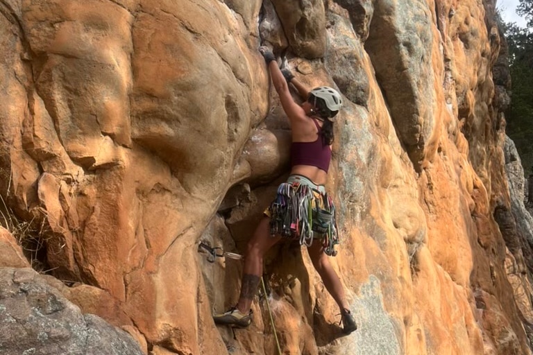 Woman in maroon sports bra climbing orange rock wall outdoors at Arapiles, reaching for hand hold, POV: behind