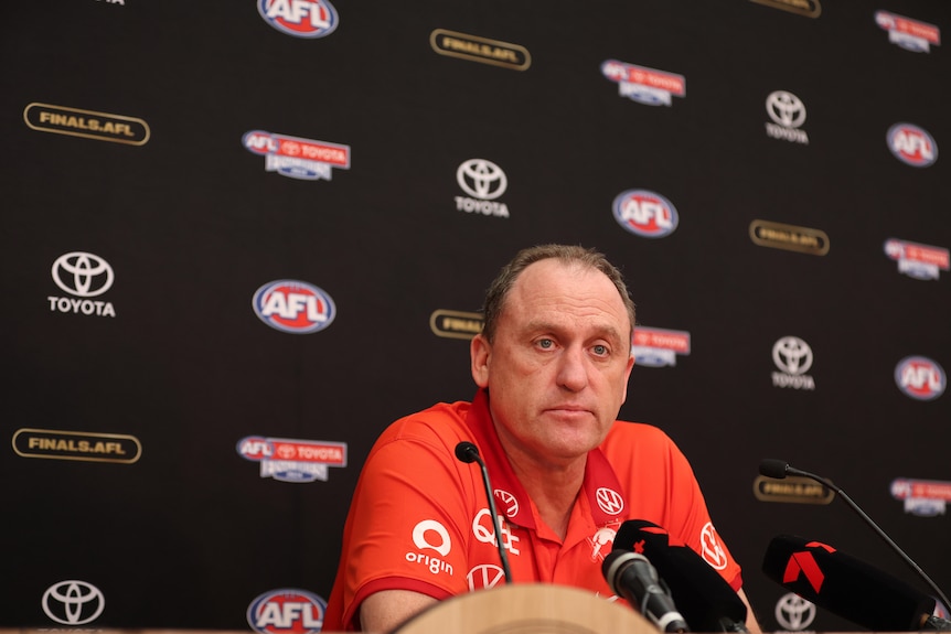 A Sydney Swans AFL coach sits at a desk looking sad as he takes part in a press conference after a grand final.