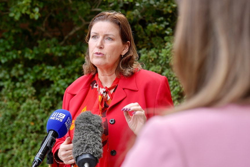 A woman wears a red blazer speaks to media. 
