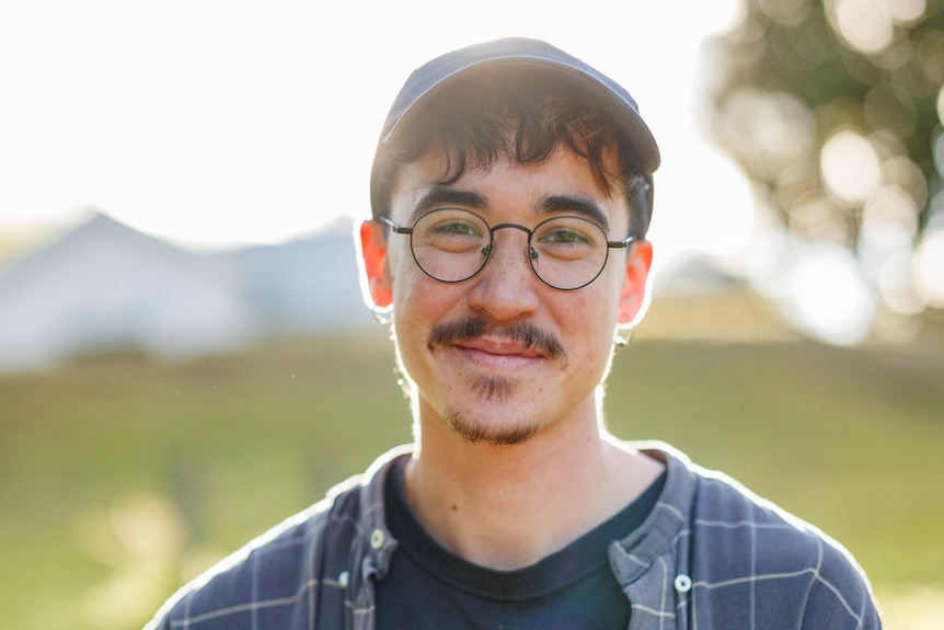 Lee Lai, with a moustache and goatee, wearing glasses, a cap and a checked shirt; the sun shining through a gum tree behind her.