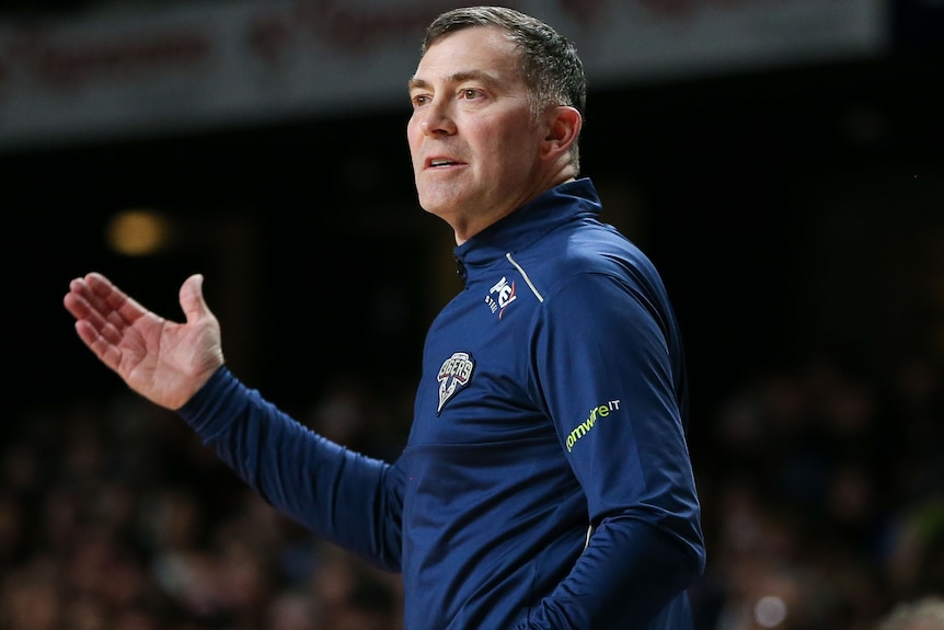 A man with short, greying hair gestures as he stands on the sidelines of a basketball game.