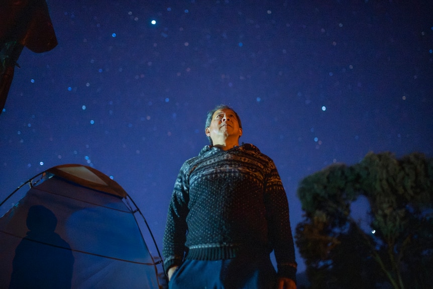 A man in a black and white patterned jumper looks up at glittery stars under an electric blue sky with a tent in background.