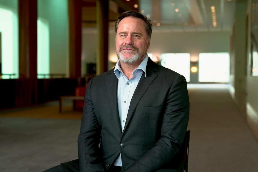 A dark haired man with a beard in a dark blazer and light blue collared shirt sits on a chair staring at the camera.
