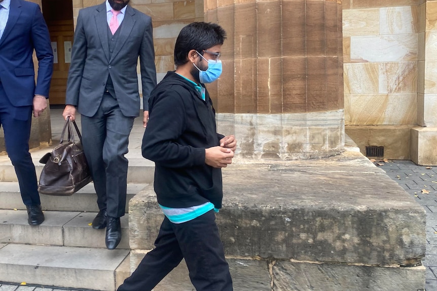 A man in a dark hoodie, glasses and face mask walks next to two men in business suits outside the court building