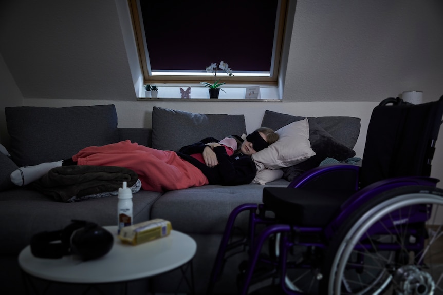 A person is on the couch, eyes covered, sleeping next to a wheelchair.