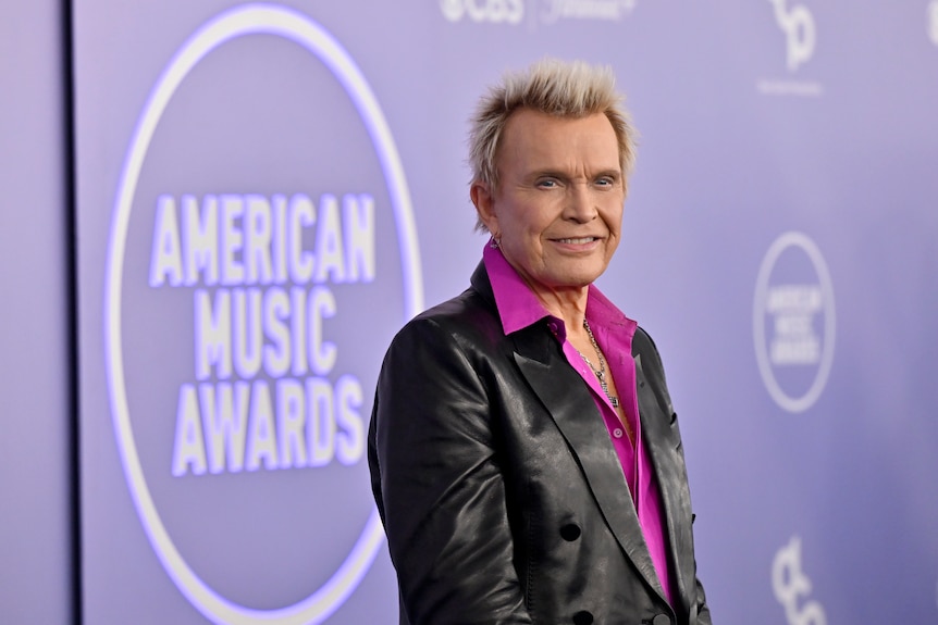 Billy Idol in black leather and a purple shirt poses.