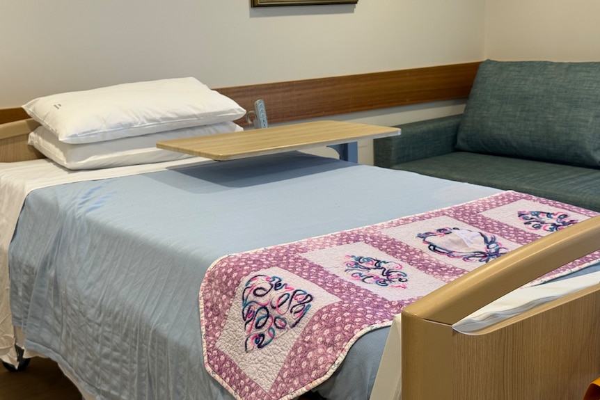 A bed with light blue sheets, a small purple comforter and a moveable overbed table is set against a wall.