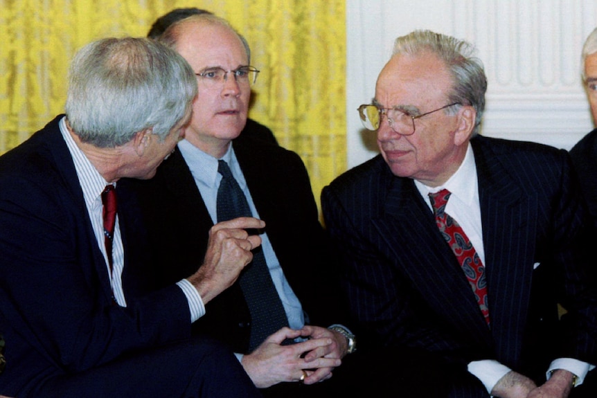 Three men sit side-by-side. The two on the outer lean over the man in the middle to speak