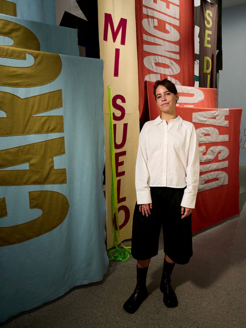 A young woman with short dark hair, wearing a white shirt and black pants, standing in front of textile banners.