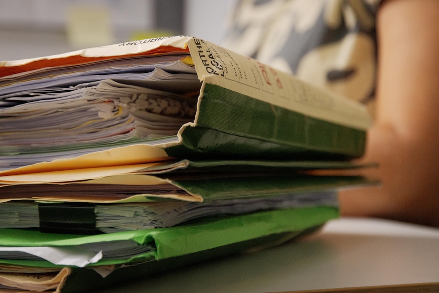 A stack of manila folders, each full of paper, on a desk, with a person in the background.