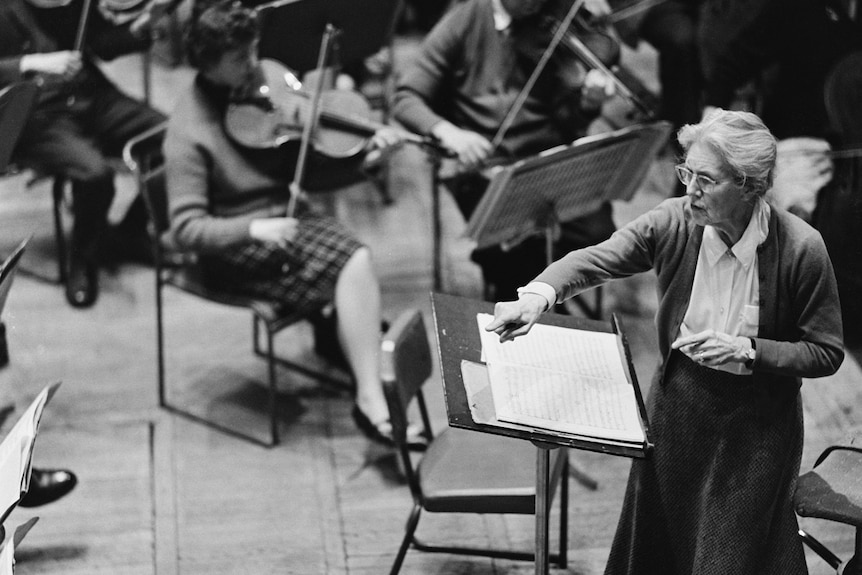 A black and white photo of Nadia Boulanger conducting an orchestra in a cardigan. She points toward the first violin section.